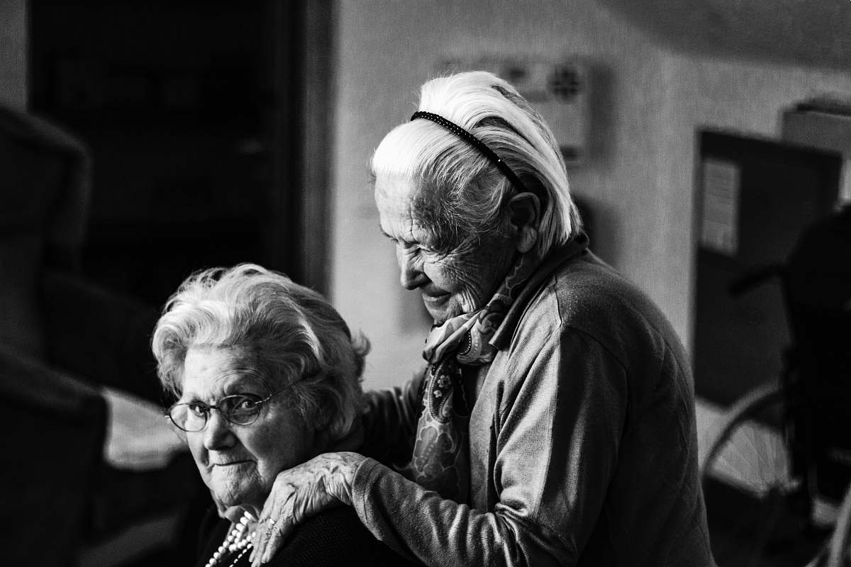 person greyscale photo of woman standing behind woman sitting on chair black-and-white