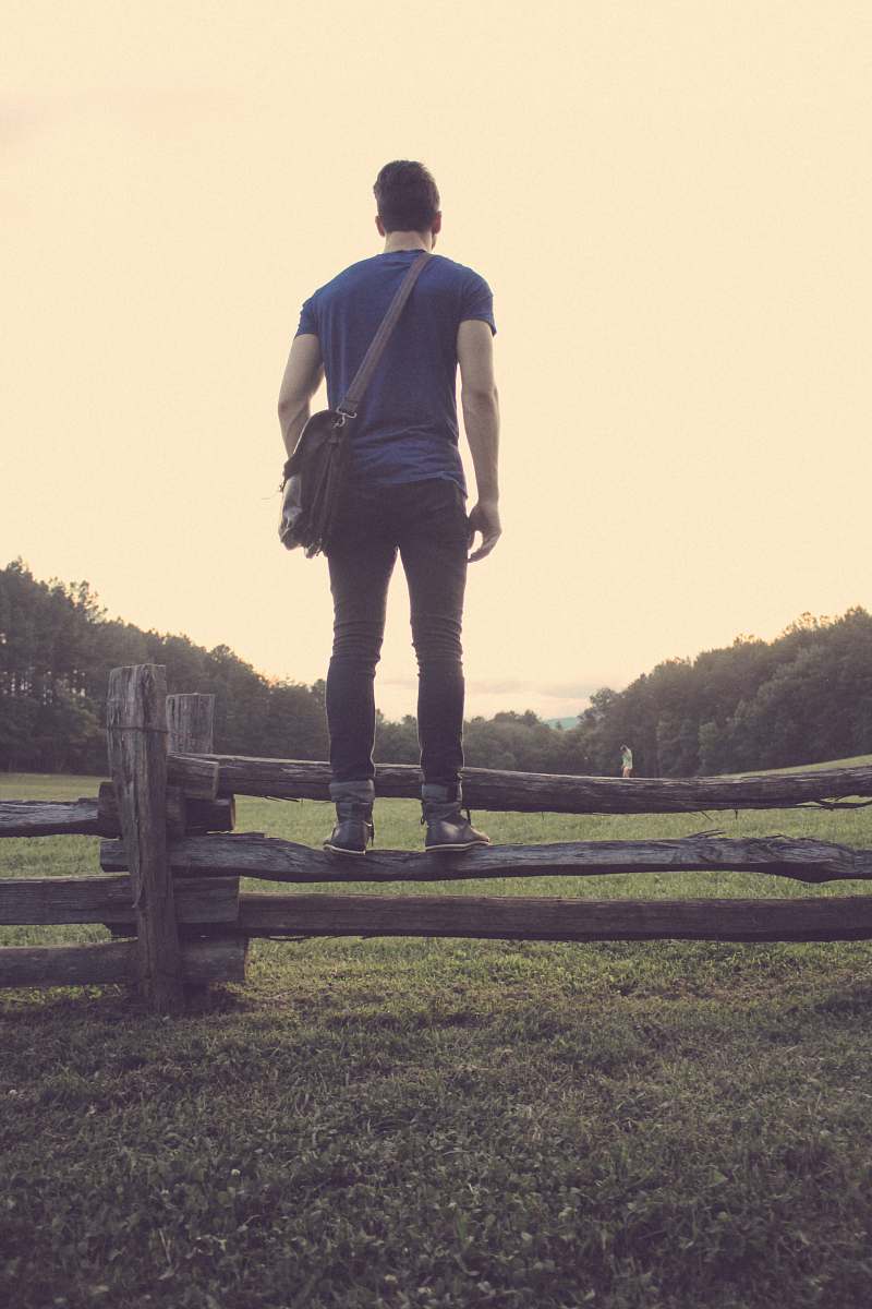 person man in blue t-shirt with black denim jeans and pair of gray high-top sneakers standing brown wooden fence during daytime human