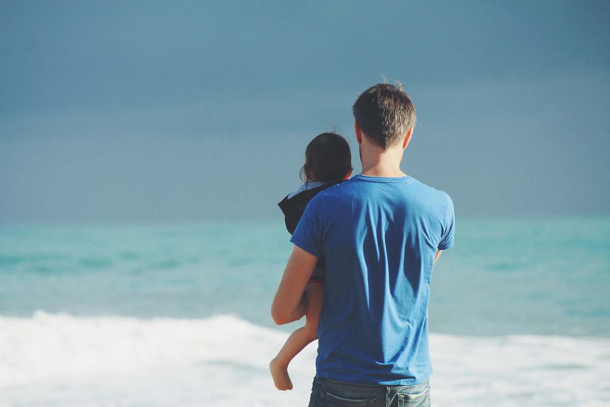 person man wears blue crew-neck t-shirt holding toddler wears black hooded jacket near ocean under blue sky at daytime father
