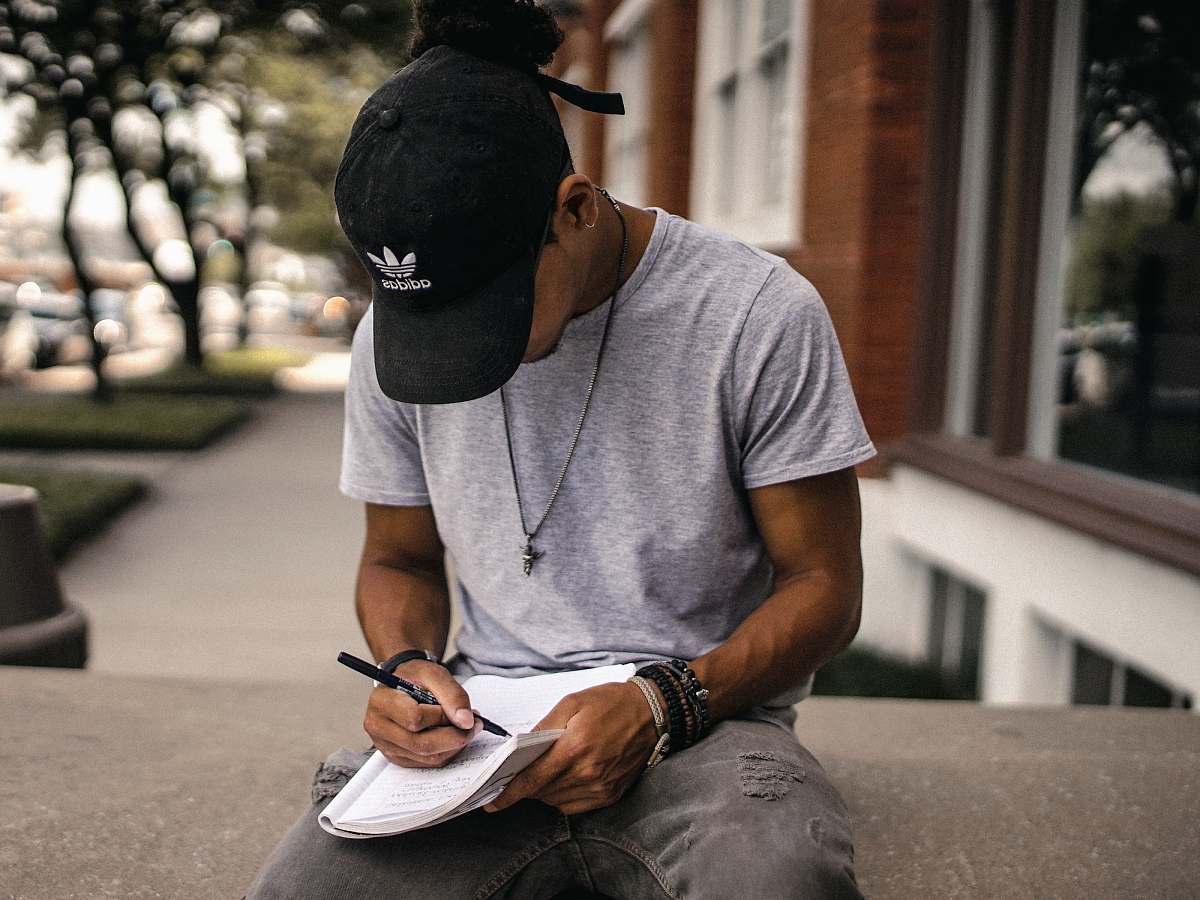 human person in black adidas cap sitting on bench writing on notebook person