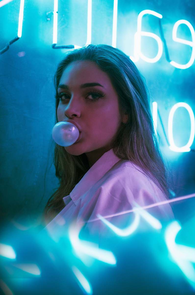 person selective focus photo of woman blowing gum standing in front of turned-on neon signage bubble