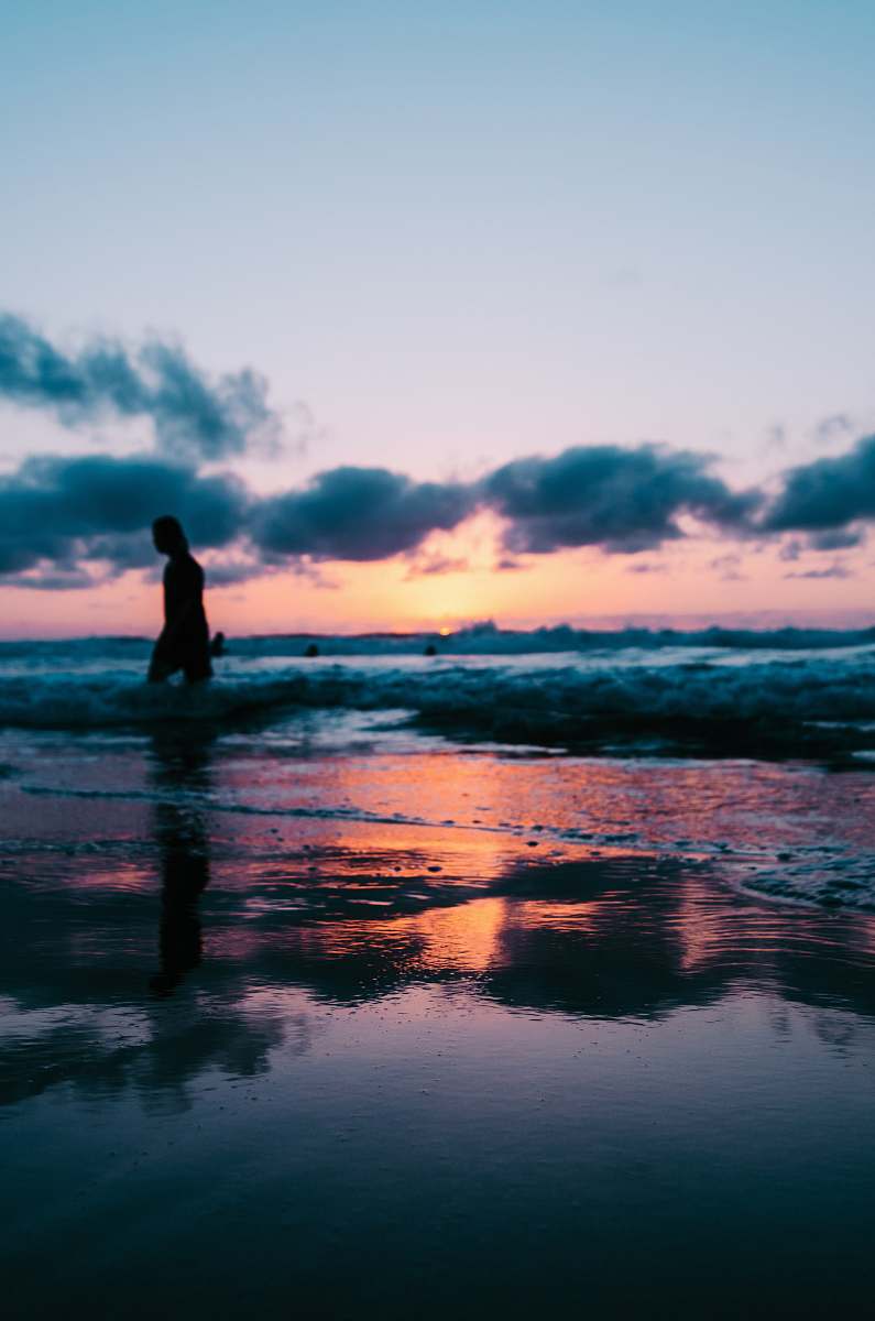 person silhouette photo of a person walking on seashore under cloudy sky during golden hour water