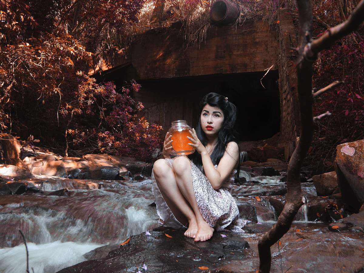 girl woman carrying glass jar while sitting on boulder beside flowing river behind brown bridge under shade of tree at daytime woman