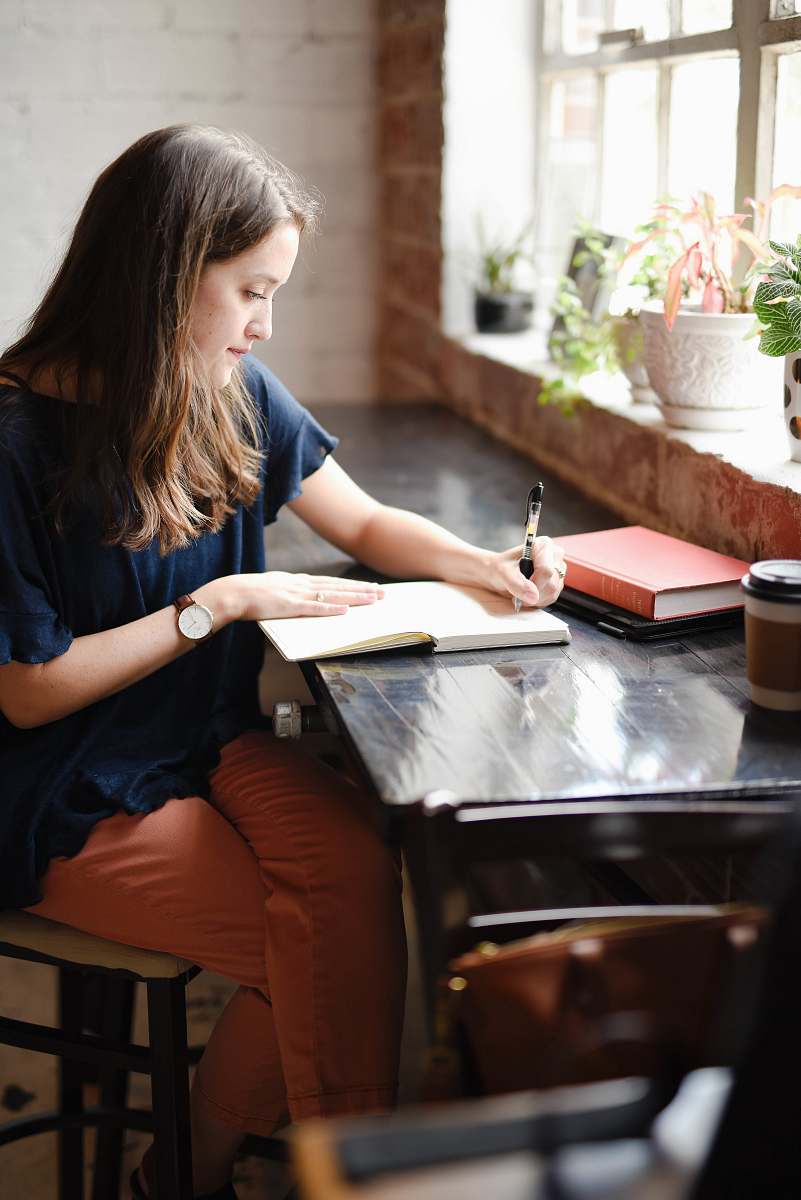 person woman sitting in front of black table writing on white book near window human