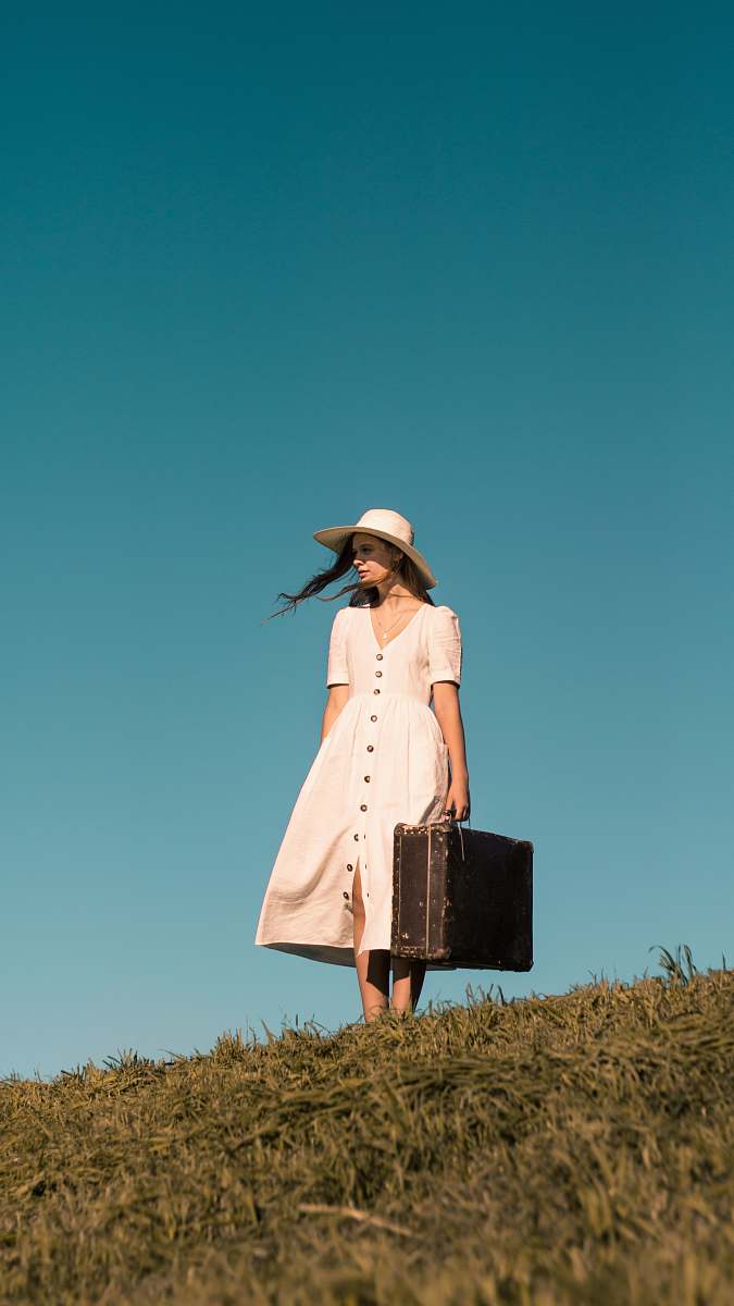 person woman standing on green grass holding brown leather suitcase wearing white button-up dress human
