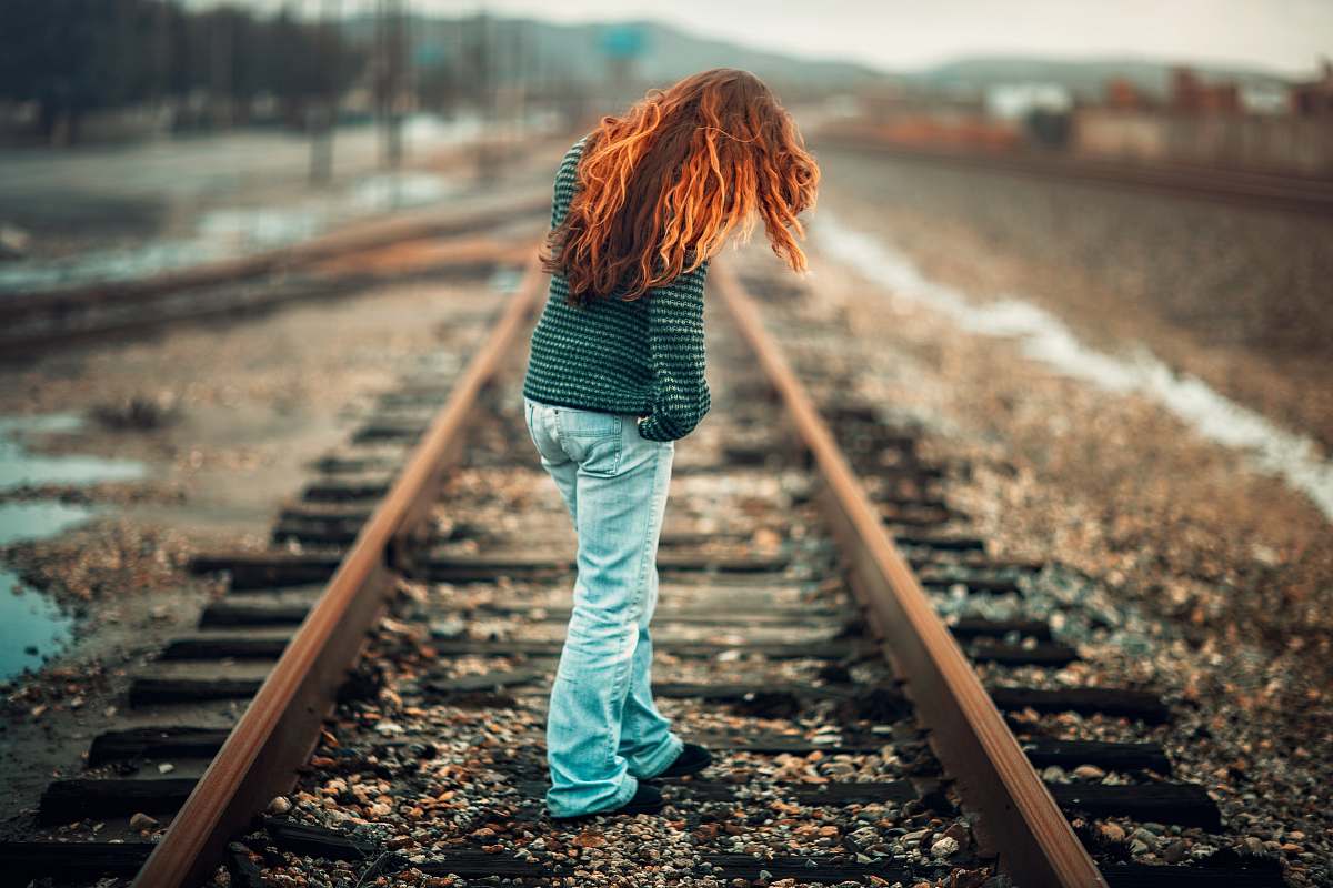 human woman with orange hair wearing black and gray stripe long-sleeve shirt and light-blue denim pants standing on train rail at day train track