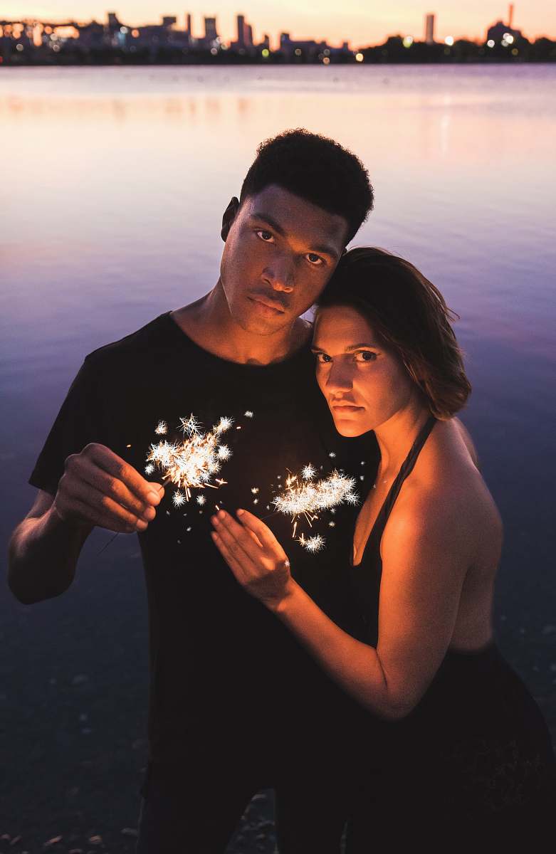 people couple holding sparklers standing near body of water at dusk human
