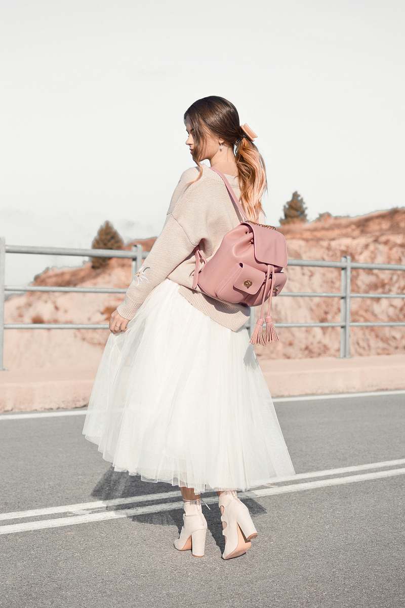 people woman standing in front of metal railings wearing brown sweater and white skirt and pair of heeled sandals carrying brown leather backpack during daytime human