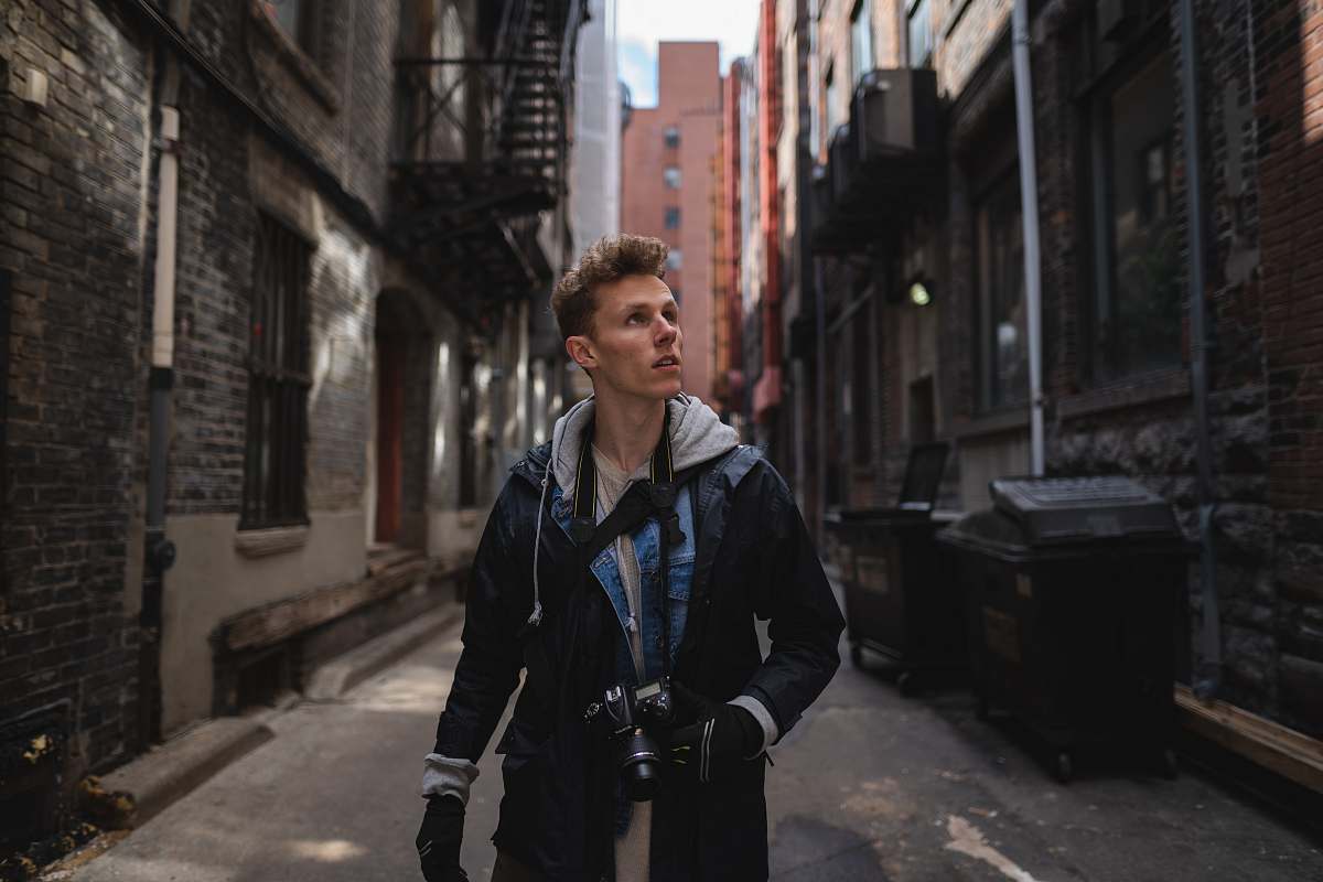 In this captivating urban scene, a man strolls along a pathway, his DSLR camera in hand, attentively looking to his right. The vibrant cityscape serves as a backdrop, showcasing dynamic street life and architecture. This image perfectly encapsulates the spirit of exploration and creativity, making it an ideal choice for travel blogs, photography websites, or marketing materials focused on urban adventure.