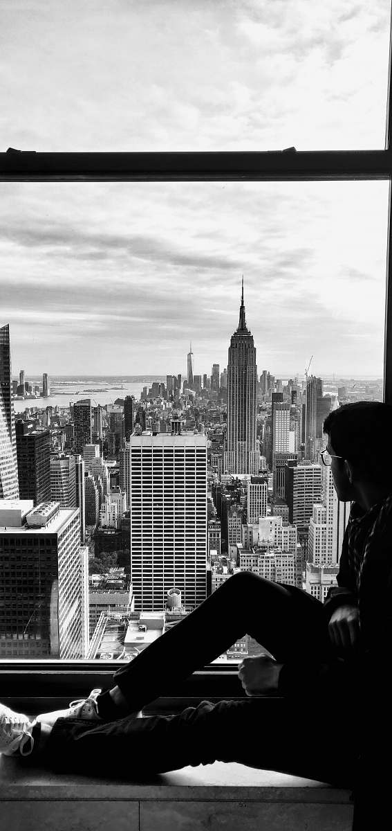 building grayscale photography of man sitting near closed window viewing New York City black-and-white