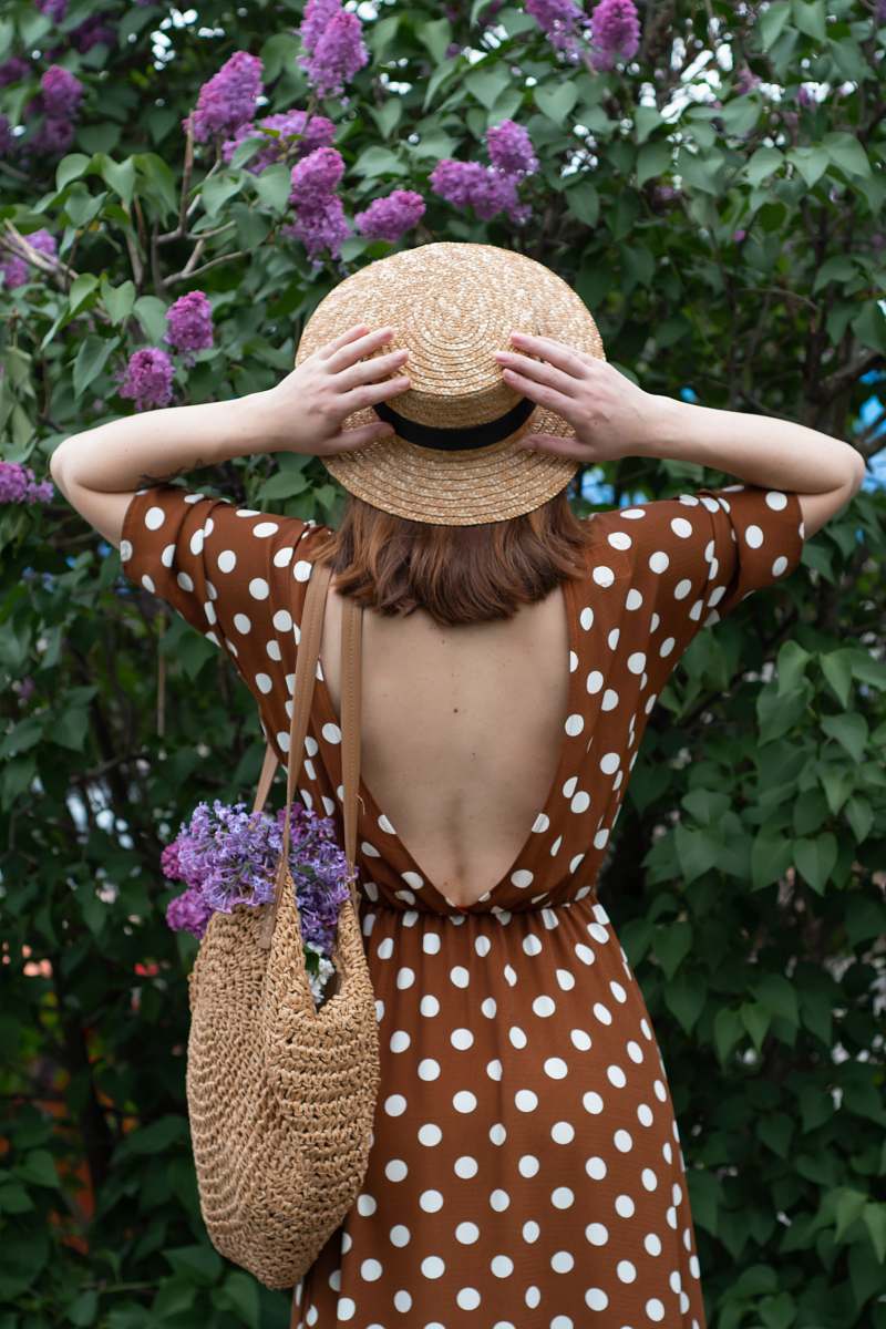 clothing woman in white and black polka dot dress wearing brown straw hat standing near purple flowers hat