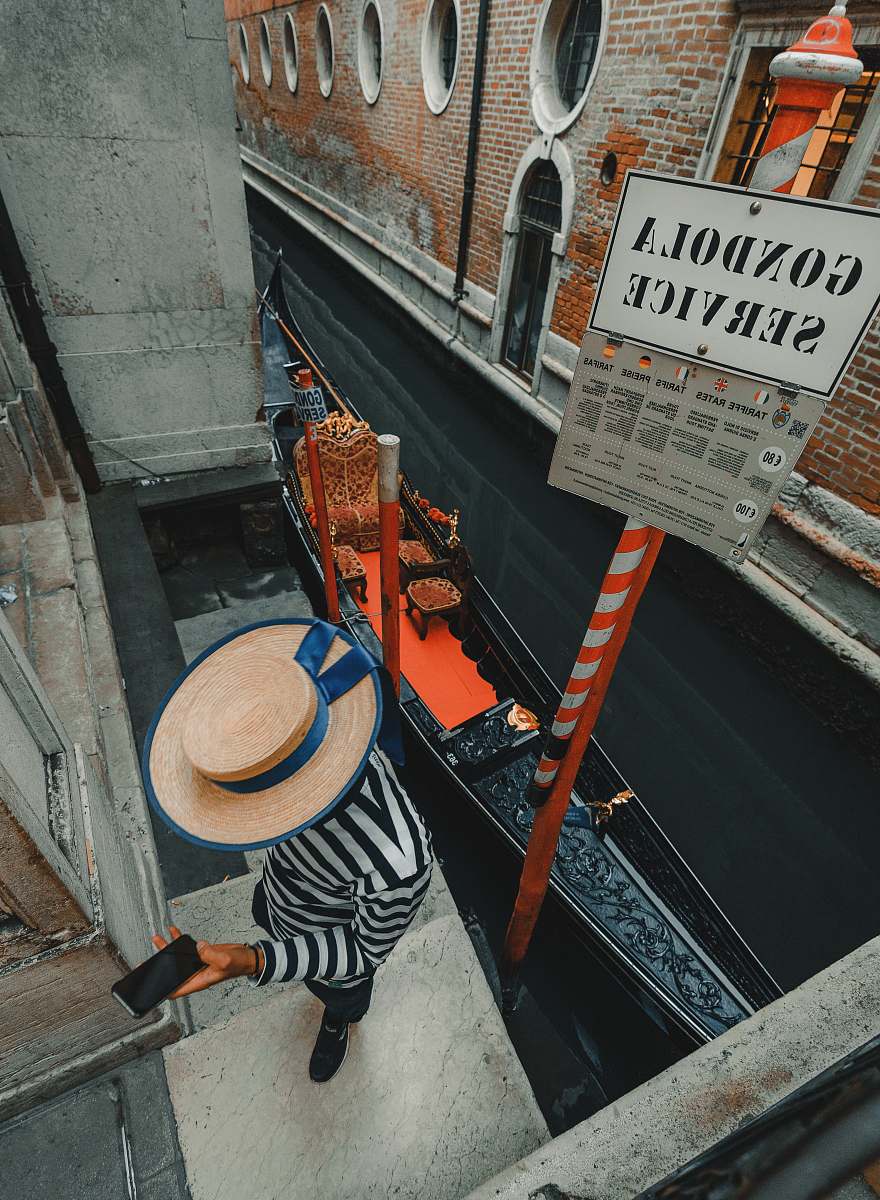 clothing person using phone while standing near Gondola beside Venice canal during daytime hat