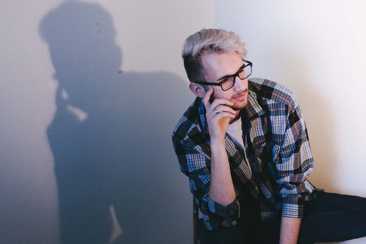 person man wearing black, blue, and red plaid collared button-up long-sleeved shirt and eyeglasses sitting and putting left hand on his face glasses