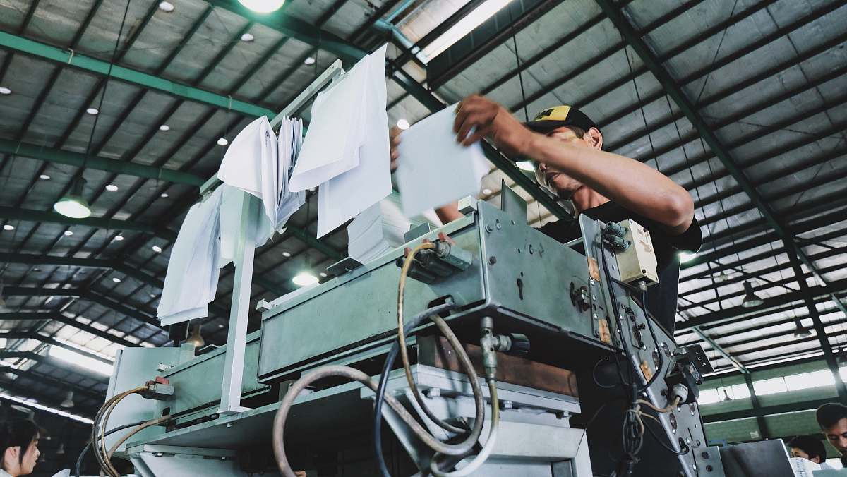 machine man holding bunch of white papers while operating large gray industrial machine inside well lighted room factory