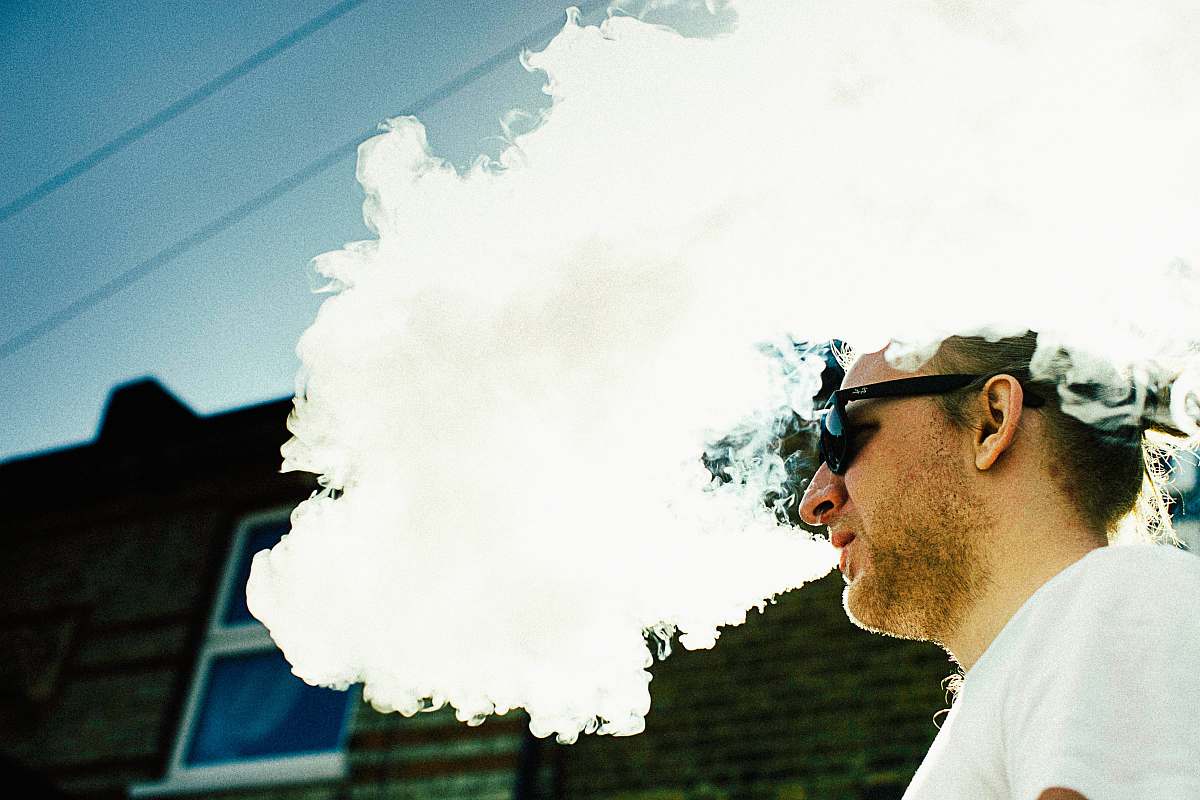 human man blowing thick white smoke on air overlooking black building wall at daytime people
