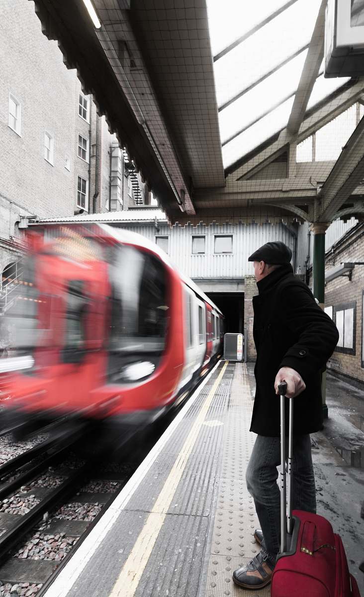 train man holding luggage waiting on railway with red train passing by people