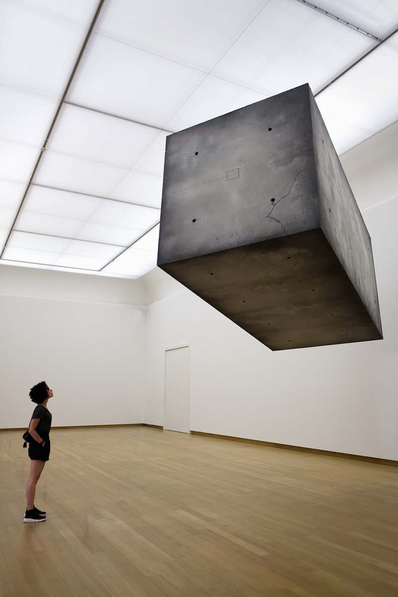 This striking image captures a woman dressed in a sleek black shirt, standing gracefully in an empty room adorned with an eye-catching sculpture suspended from the ceiling. The minimalist background emphasizes her contemplative pose, making it a perfect visual for themes of solitude, artistic expression, or modern living environments. Ideal for use in fashion, interior design, or art-related projects.