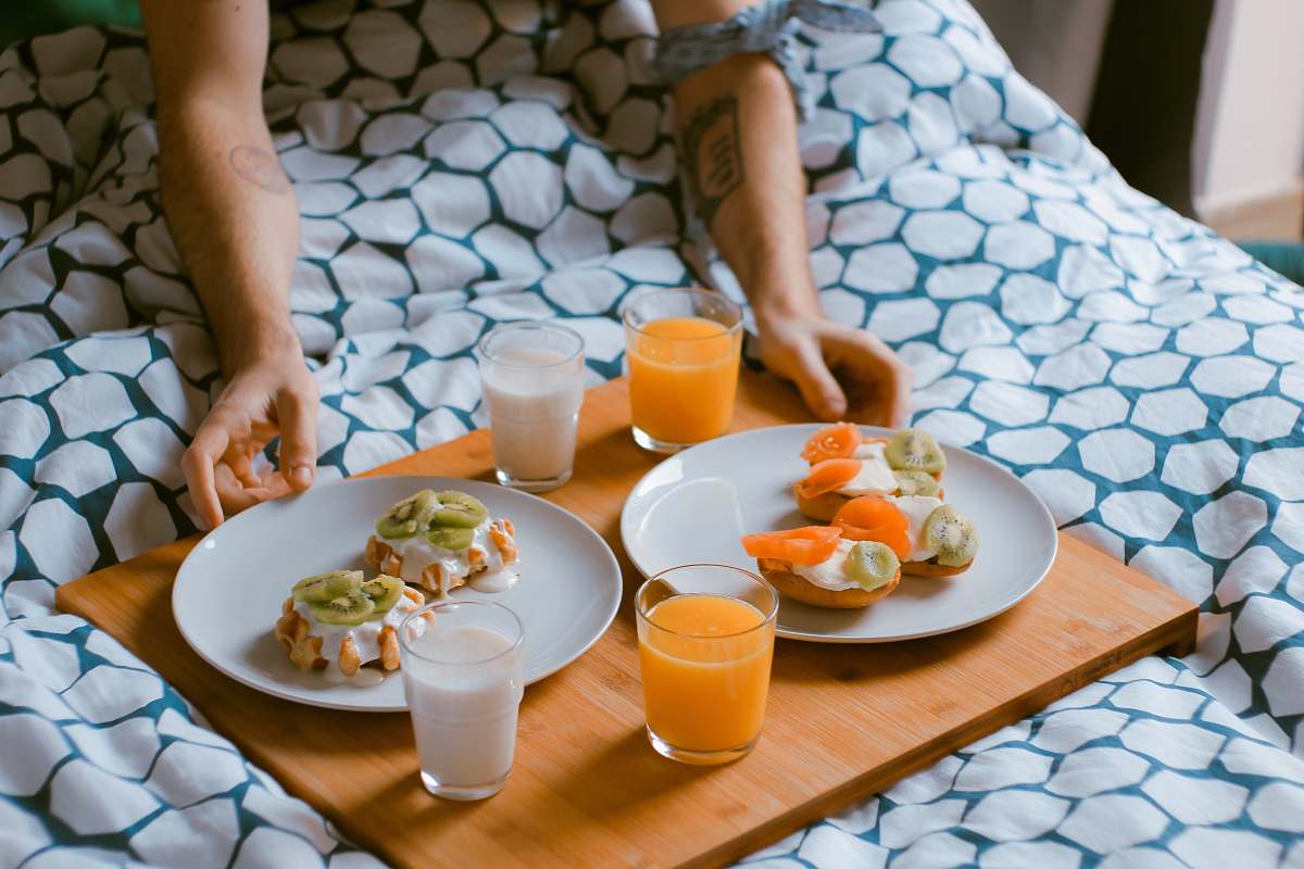 meal person serving pastries on white ceramic plates with fruit juice glasses on wooden tray on top of bed breakfast