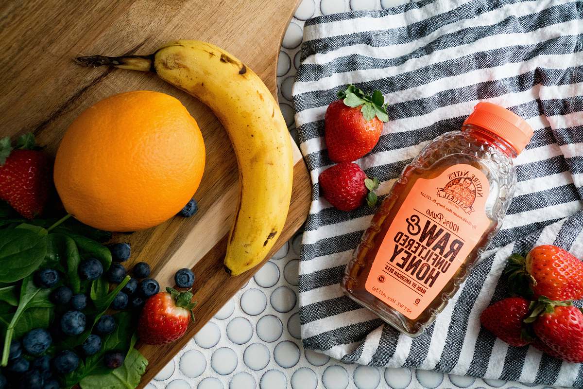fruit raw and unfiltered honey bottle between strawberries with blueberries, citrus, and banana fruits nearby blueberry
