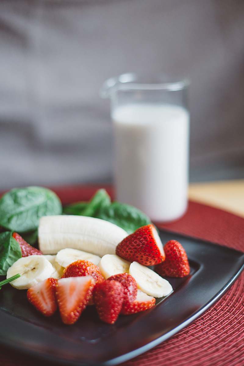 fruit sliced strawberries and bananas on black plate beside white liquid-filled clear drinking glass strawberry