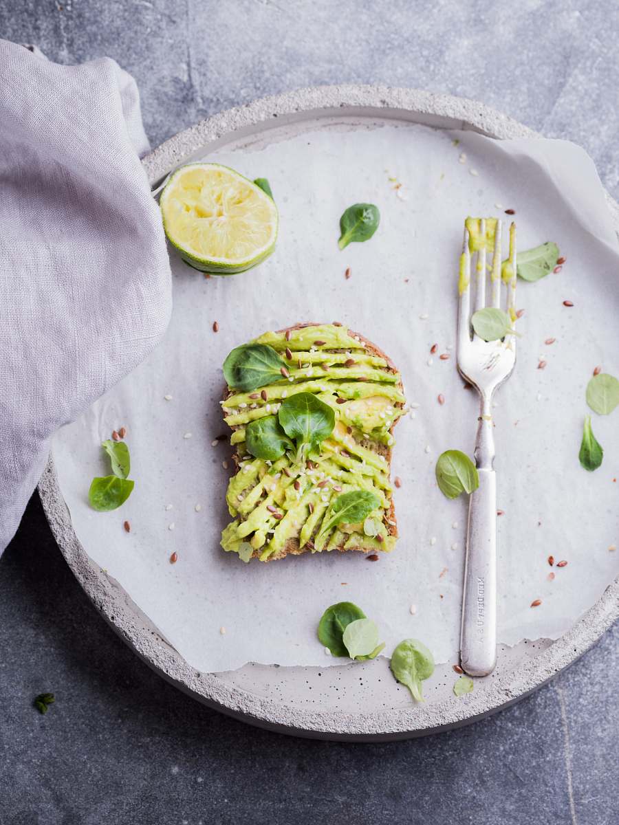 plate wheat bread with avocado spread beside white plastic spoon and pressed lime on round white plate breakfast