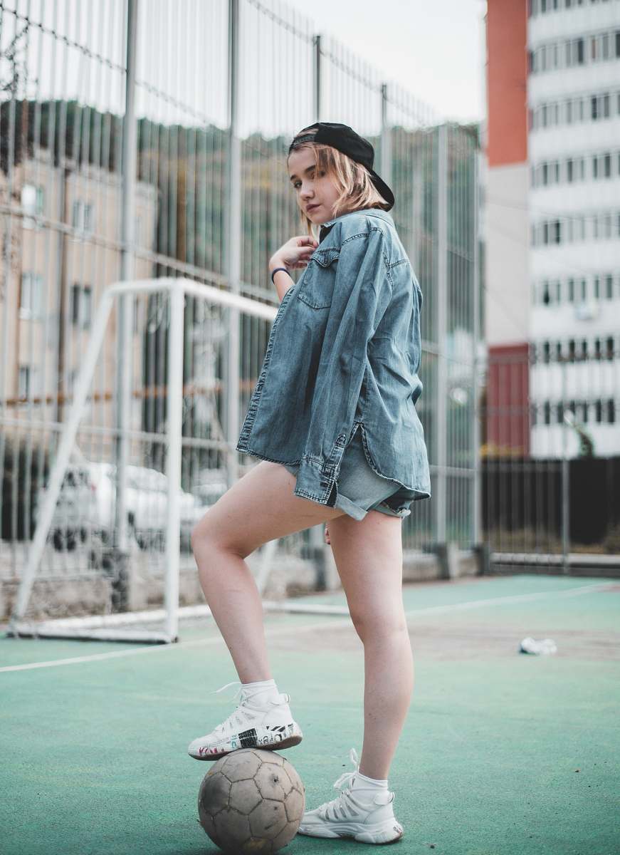 apparel woman wearing blue denim jacket standing and stepping right foot on white soccer ball in green field during daytime clothing