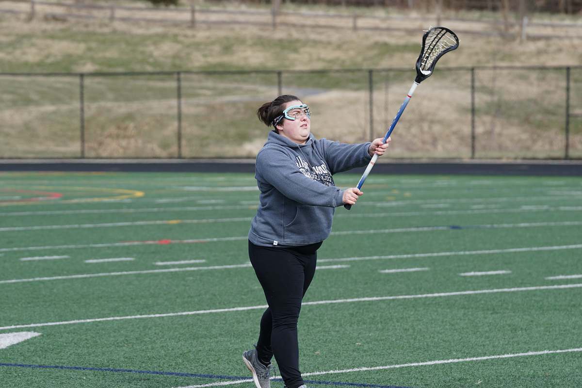 person woman wearing gray jacket standing and holding lacrosse stick on green field at daytime racket