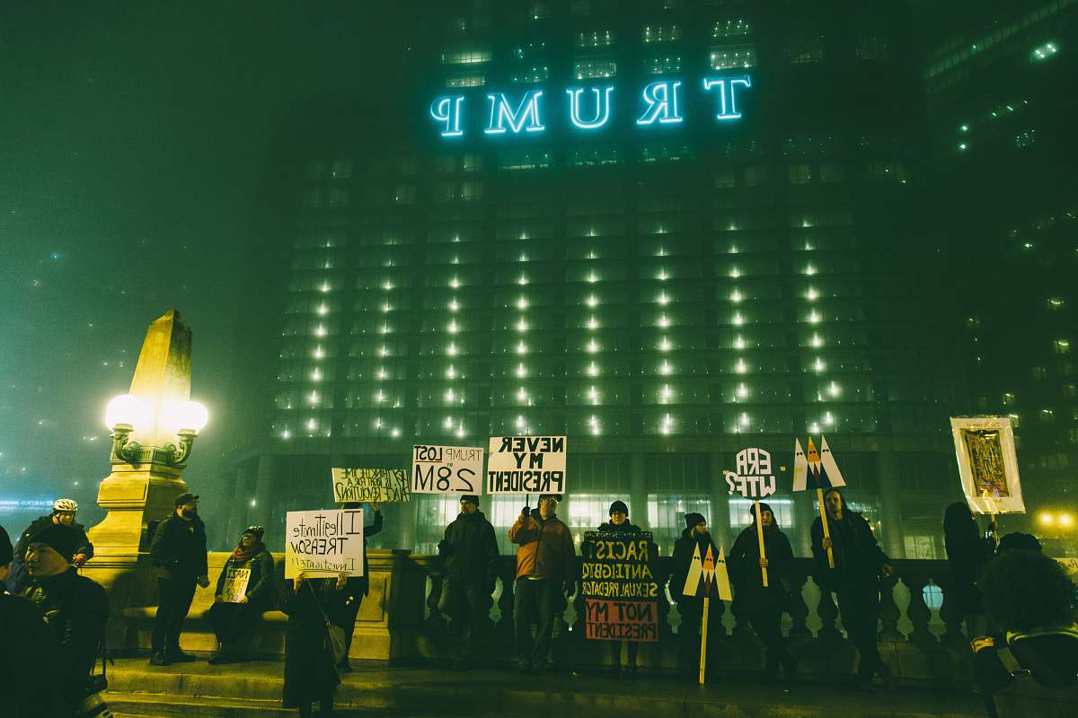 A striking image captures a solitary figure standing on a bridge, framed by a modern building in the background, as a group of passionate protesters gather below. This evocative scene showcases the energy of civic engagement, making it ideal for news articles, social media posts, and campaigns focused on activism, social justice, or urban life. The contrast between the stillness of the individual and the dynamic crowd below adds depth to the narrative of community action.