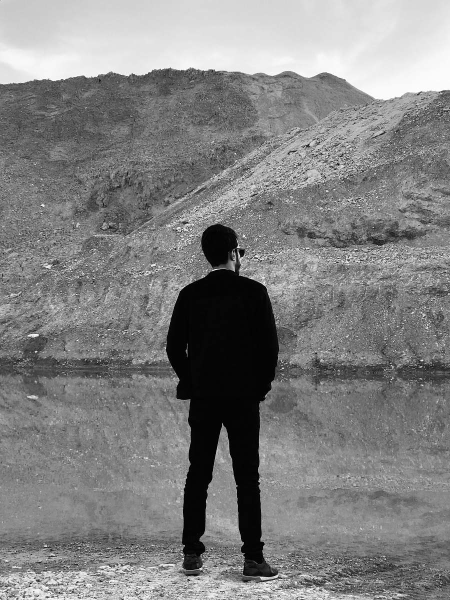 standing grayscale photo of man standing in front of body of water and mountain human