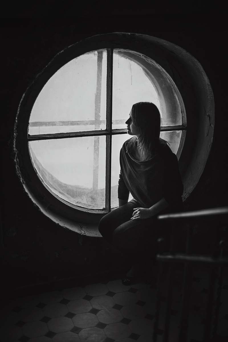 window grayscale photo of woman sits near round wooden framed window person