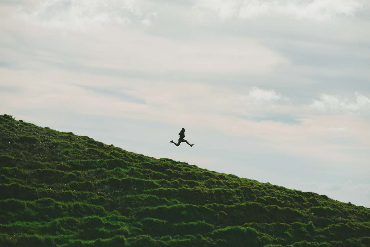 hill person jumping on grass field under cloudy sky during daytime jumping