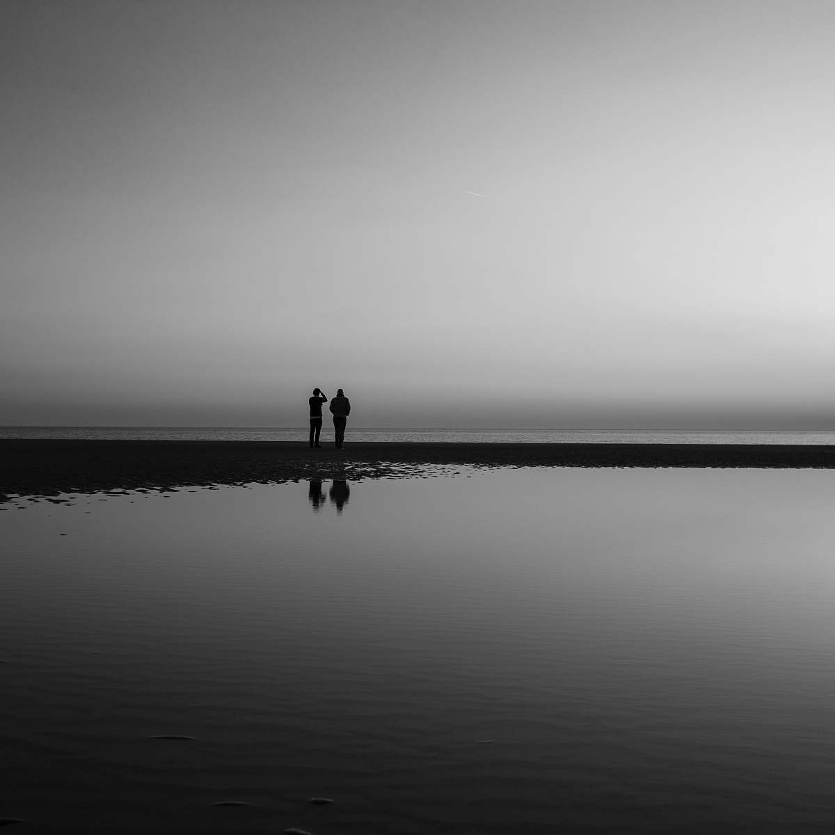 person silhouette of two person standing beside calm water black-and-white