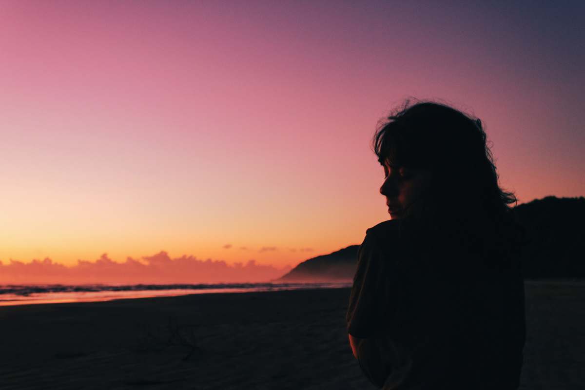 Capture the tranquility of nature with this stunning image of a woman standing on the beach at sunset, gazing thoughtfully to her right. The warm hues of the setting sun cast a magical glow, enhancing the peaceful ambiance of the scene. This versatile photograph is perfect for lifestyle blogs, wellness articles, and travel websites, evoking feelings of calmness, reflection, and connection with nature.