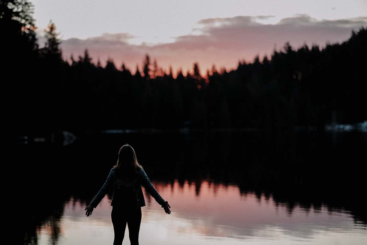 person woman standing with hands spread on side facing lake at sundown silhouette