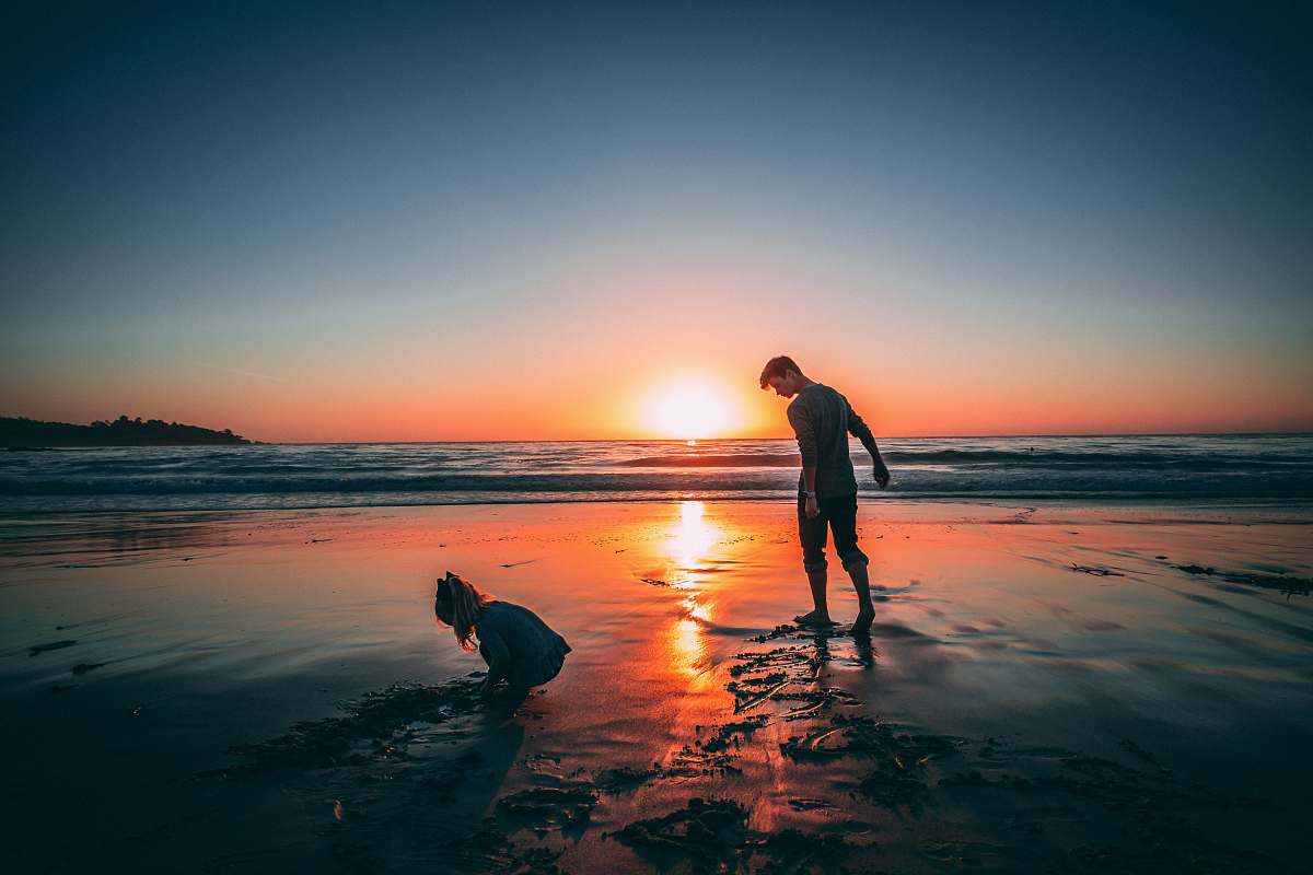 human man standing on beach beside girl playing sand during sunset person