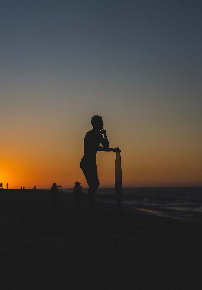 outdoors person standing and holding surfboard on shore during golden hour human
