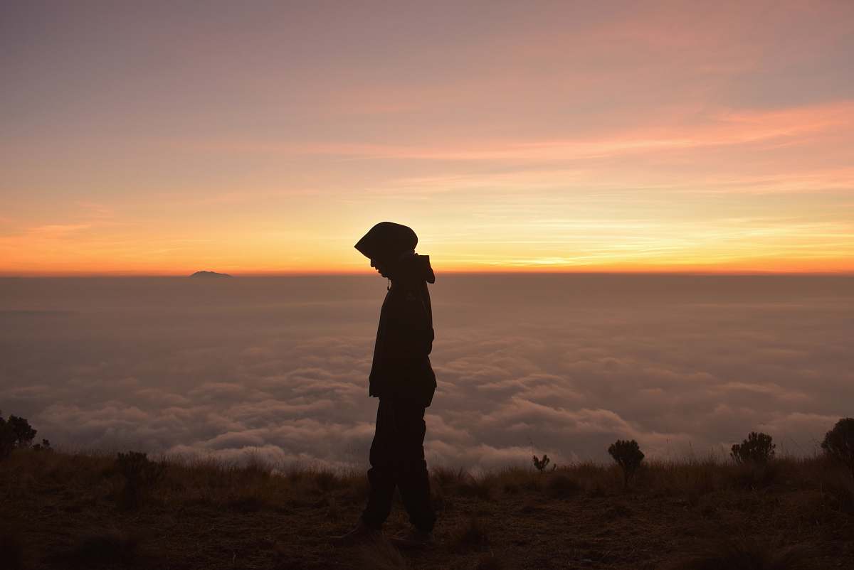 human silhouette of person wearing hoodie standing near cliff with clouds ahead during golden hour people