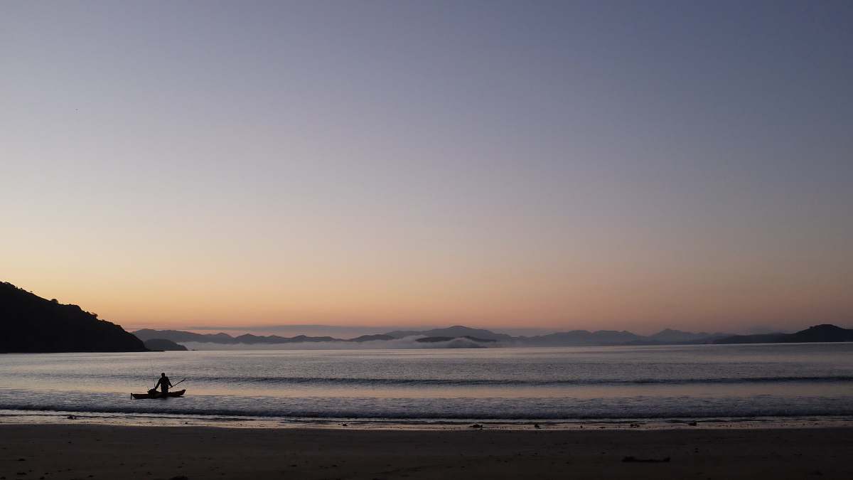 beach silhouette photo of person riding on a boy on open sea near mountains during golden hour coast