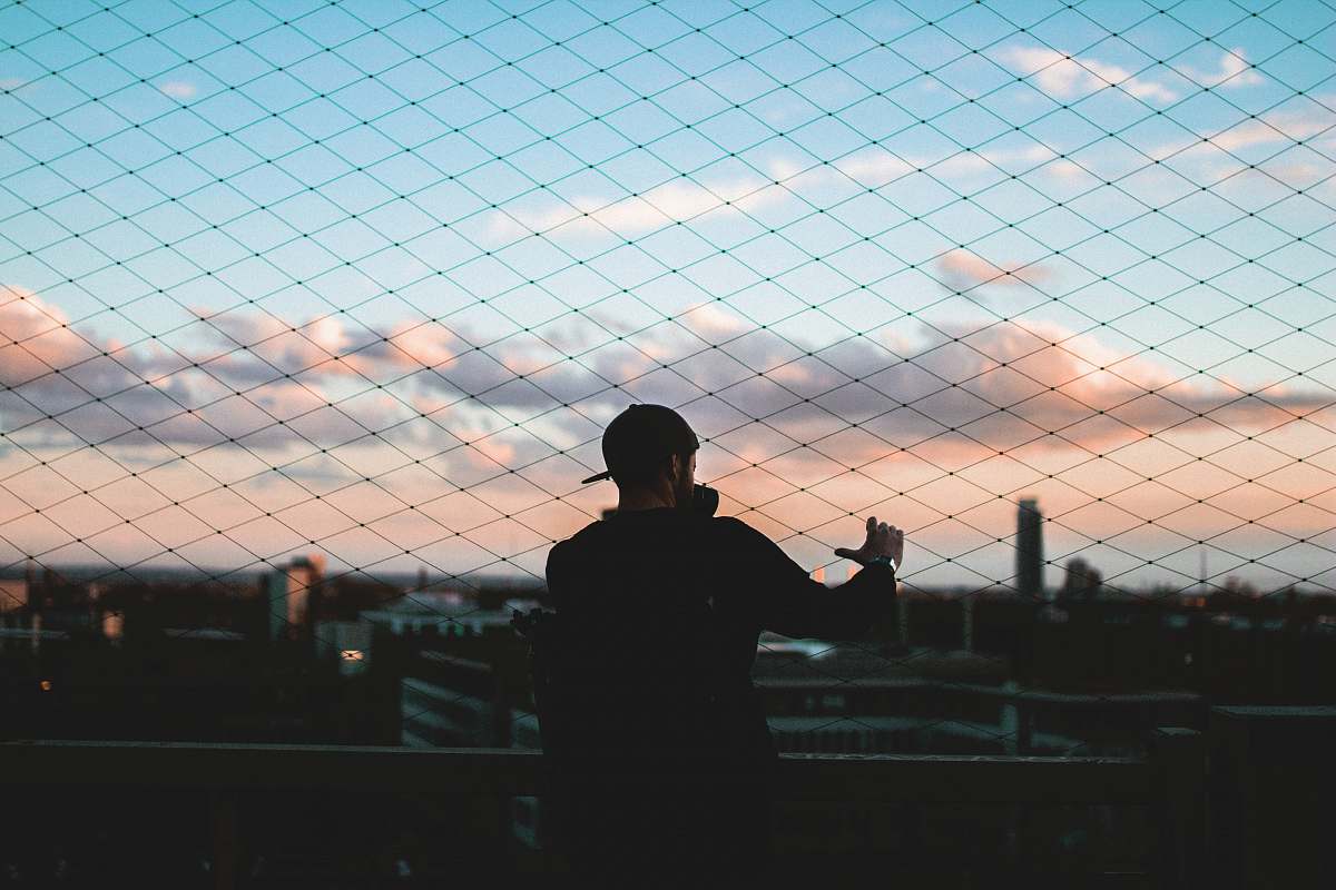 person man standing behind chain link fence holding to fence during golden hour skyline