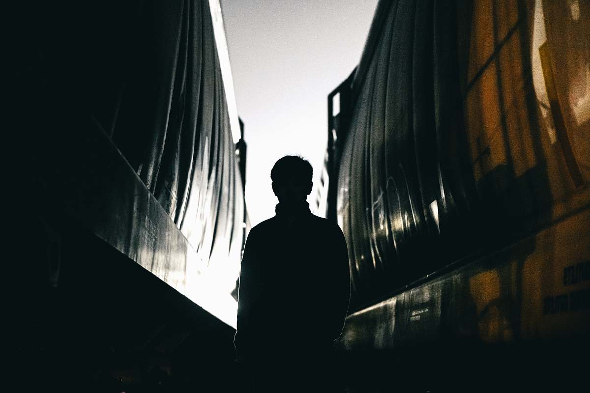 This captivating image captures a lone figure standing in a dimly lit gap between two towering buildings, silhouetted against the backdrop of a bustling urban environment. The scene conveys a sense of intrigue and solitude, as the trains pass by, highlighting the contrast between the dynamic movement of city life and the stillness of the man's presence. Ideal for use in urban lifestyle blogs, travel articles, or any project that explores themes of solitude, mystery, or modern cityscapes.