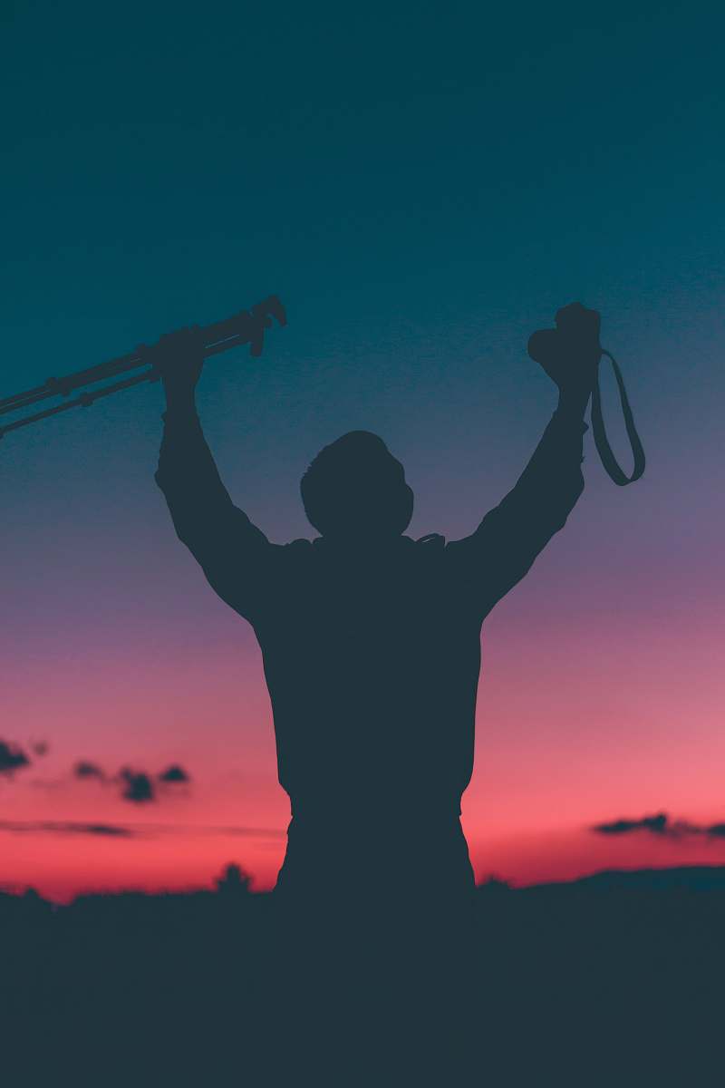 dusk silhouette of a person raising camera and tripod with pink and blue sky sunset