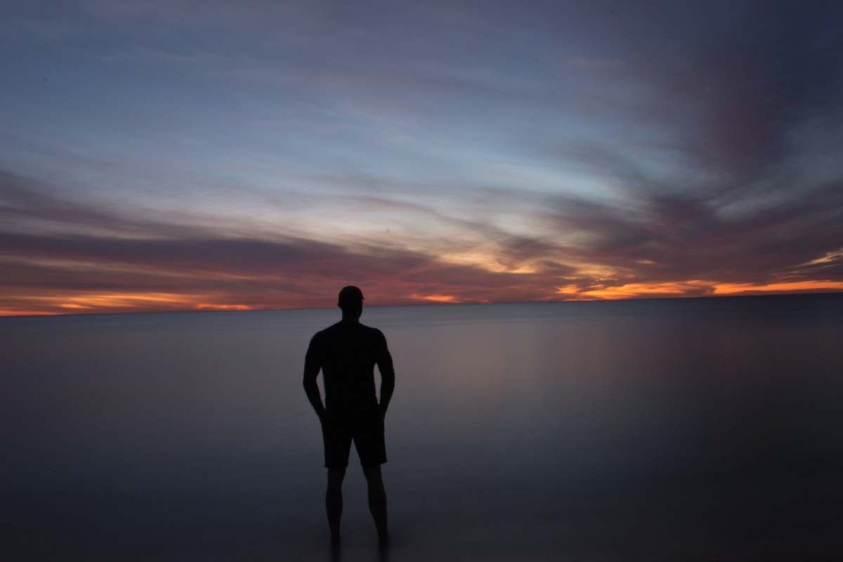 people silhouette of person standing near calm body of water during golden hour person