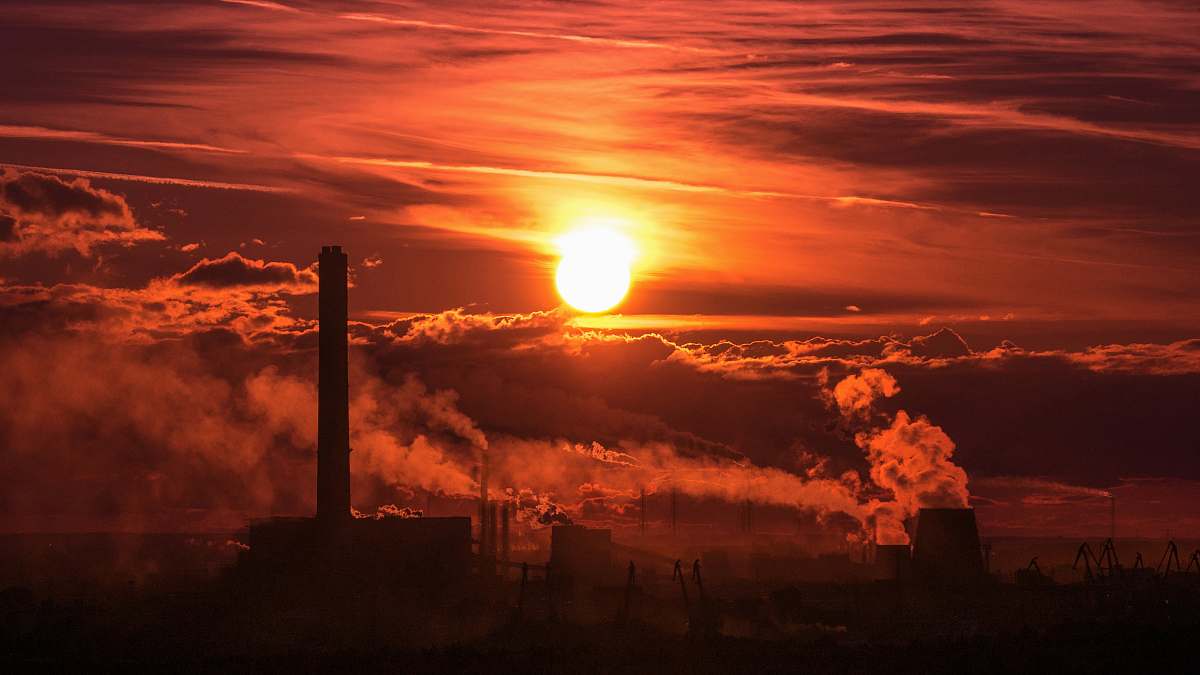 Capture the breathtaking beauty of a sunset illuminating a tall building and a large factory adorned with smoke stacks. This striking scene features vibrant orange clouds that frame the industrial landscape, creating a powerful contrast between nature and urban development. Perfect for use in environmental discussions, industrial brochures, or as a striking backdrop for corporate presentations, this image embodies the harmony and tension between industry and the natural world.