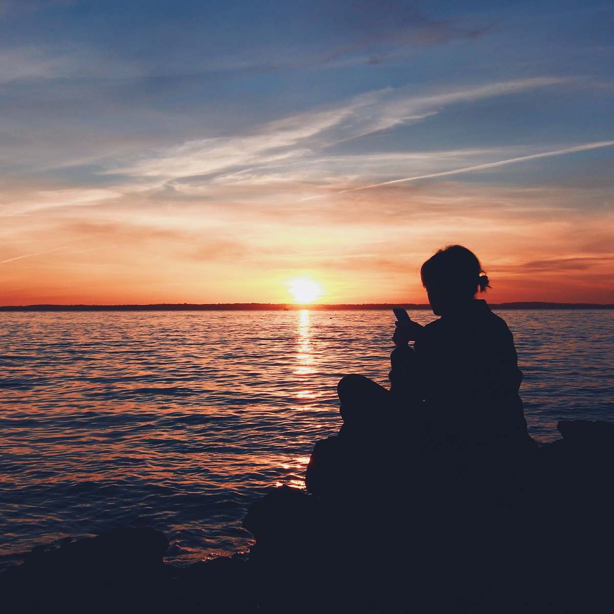 human silhouette photo of person sitting on boulder near body of water person