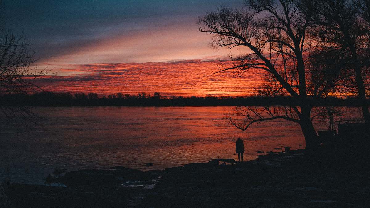 sunrise silhouette of person standing near tree and body of water at nighttime sunset