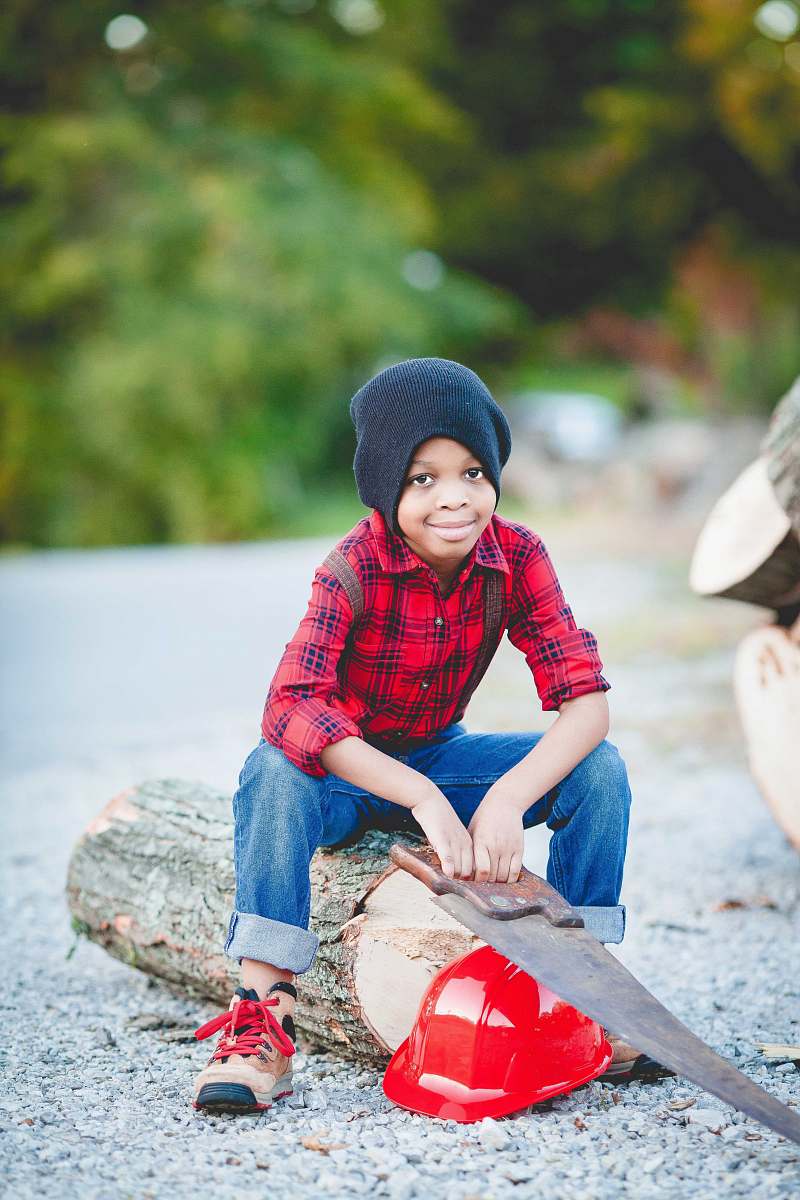 people boy sitting on log holding handsaw over red hard hat person