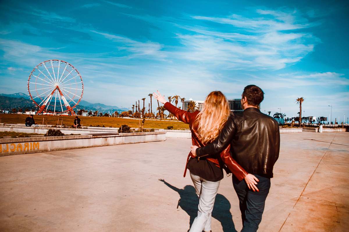 people couple walking girl pointing red ferris wheel under blue sky and white clouds person