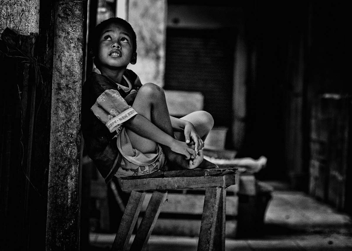 person grayscale photo of boy sitting on wooden bench beside wall while looking up black-and-white