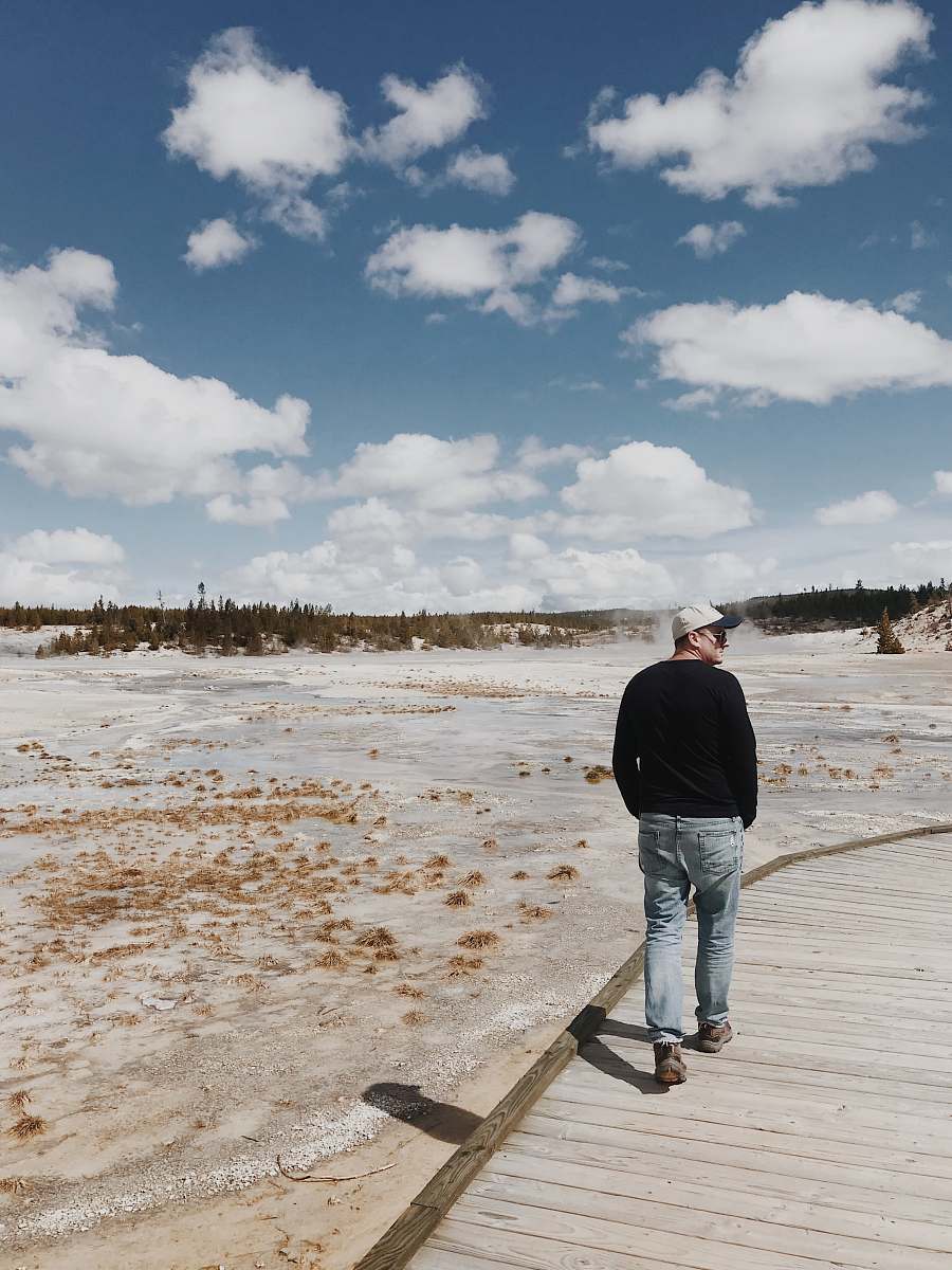 people man in black sweatshirt and blue denim jeans walking on wooden pathway under blue and white skies during daytie person