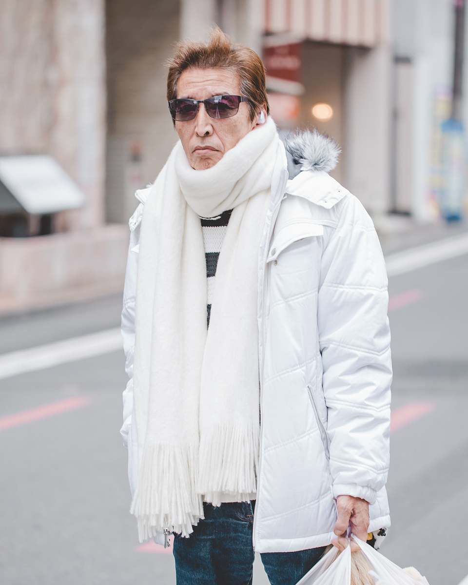 Capture the essence of winter fashion with this striking image of a man in a white parka jacket and a white scarf, strolling elegantly on a vibrant street during the day. Accompanying him is a woman dressed in a chic white coat and scarf, embodying a modern and sophisticated style. This scene is perfect for use in lifestyle blogs, fashion articles, or marketing campaigns that aim to convey elegance and urban charm.